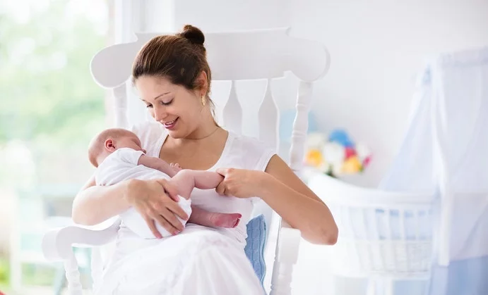 young mother holding her newborn child. mom nursing baby. woman and new born boy relax in a white bedroom with rocking chair and blue crib. nursery interior. mother breast feeding baby. family at home